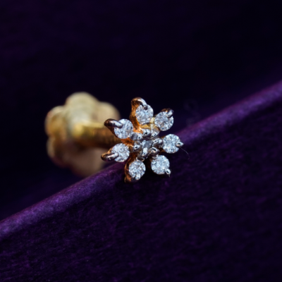 A gold nose pin with a flower-shaped design, featuring multiple diamond stones, placed on a purple cushion.